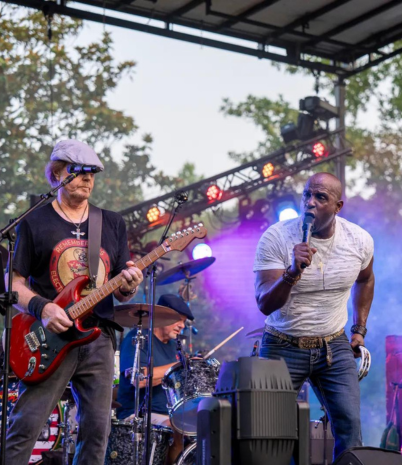 A band performs at a festival. Shown are a man playing guitar and a male lead singer.